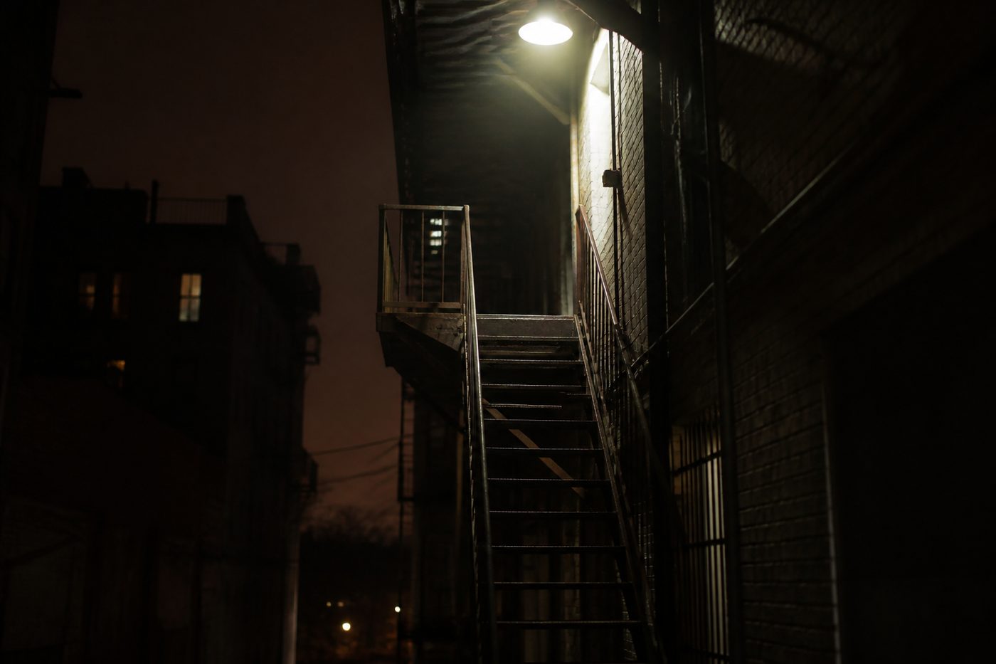 Metal fire escape stairway in a narrow alleyway lit by a single overhead light at night